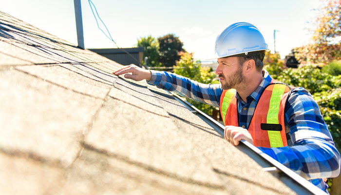 man with hard hat standing on steps inspecting house roof for repair